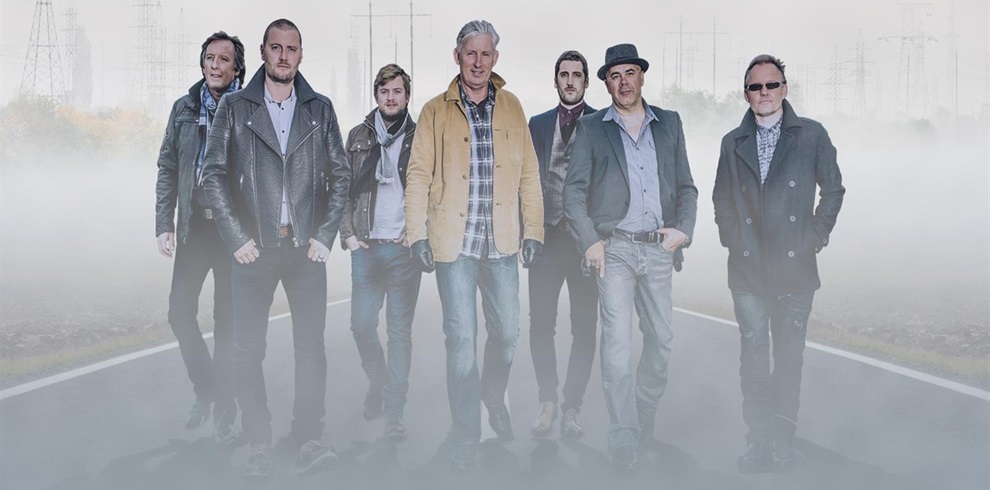 A group of eight people walking down a foggy road, with power lines and towers visible in the background. The individuals are dressed in casual clothing such as jackets, jeans, and hats, and their faces are blurred.