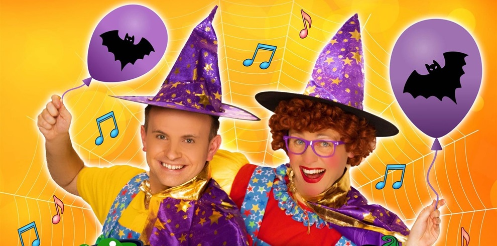 Two people in colorful costumes wearing purple wizard hats with yellow stars, holding purple balloons decorated with black bat silhouettes. Their faces are blurred. The background is bright orange with a large spider web design and scattered musical notes.