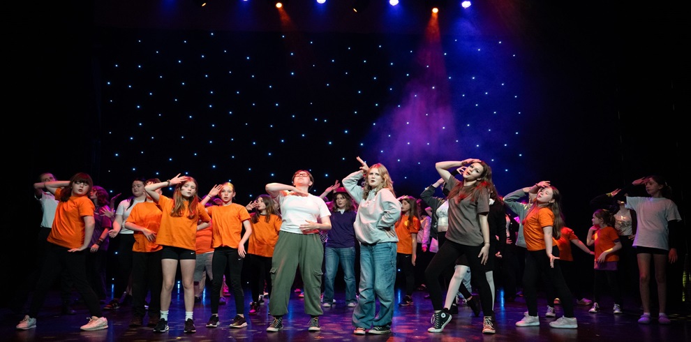 A large group of young performers is mid-dance on a theatre stage, striking dramatic poses with one hand saluting on their foreheads. The stage is lit with vibrant blue, orange, and purple spotlights, and the backdrop features a curtain of twinkling star-like lights. Many of the performers wear casual clothing in orange, white, grey, and black, creating a colorful and energetic scene. The atmosphere is lively and theatrical, suggesting a musical theatre performance in progress.