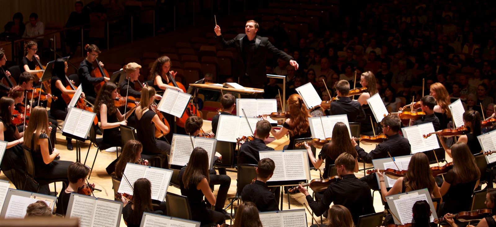 A large school symphony orchestra is performing on stage in a concert hall. The conductor, dressed in black, stands at the center on a podium with both arms raised, leading the musicians. The orchestra consists of students playing string instruments such as violins, violas, and cellos, all reading from sheet music. The musicians are dressed in formal black attire. The stage is brightly lit, while the audience in the background is dimly visible in the shadows.
