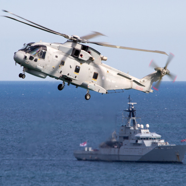 Royal Navy Merlin helicopter in flight above navy ship in the sea.