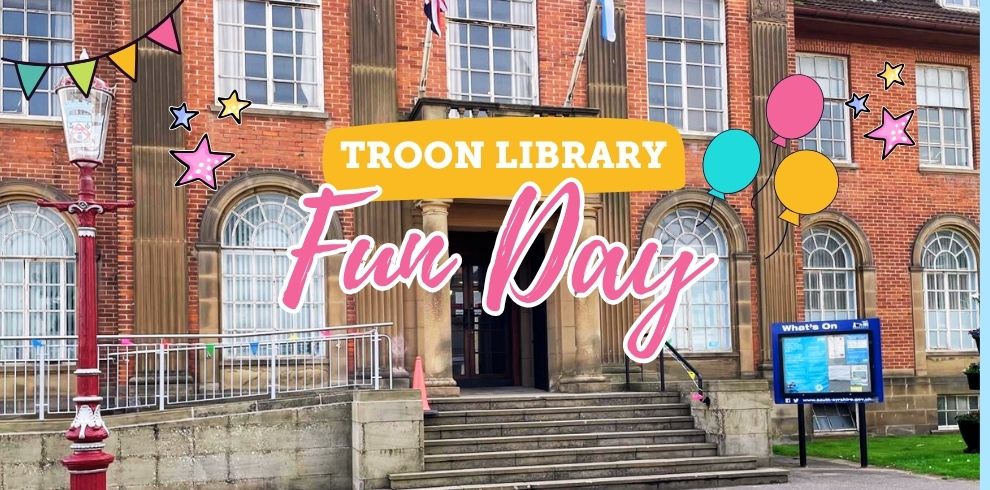A promotional image for "Troon Library Fun Day" set in front of the historic Troon Municipal Buildings. The text "Troon Library Fun Day" is boldly displayed in colorful, playful fonts. The background shows a red-brick building with arched windows and a columned entrance. Decorations include illustrated balloons, bunting, and stars, creating a festive and inviting atmosphere. A blue "What’s On" events board is visible in the lower right corner near the steps.