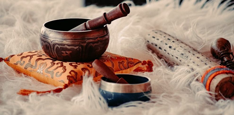 A Tibetan singing bowl with a wooden mallet rests on an ornate orange cushion, placed on a fluffy white surface. Nearby are other ritual instruments, including another small bowl, a cylindrical shaker with holes, and a beaded stick wrapped with orange cord.