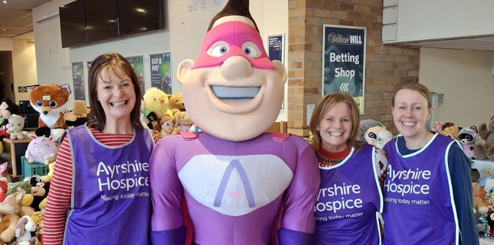 Three smiling women wearing purple 'Ayrshire Hospice' volunteer vests stand beside a person in a large pink and purple superhero costume with a big grin and a stylized 'A' on their chest. Behind them is a table filled with colorful stuffed animals, and a 'William Hill Betting Shop' sign is visible on the wall.