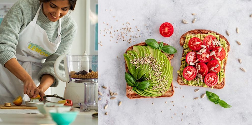A woman wearing a white apron prepares food in a kitchen, slicing ingredients beside a food processor. Next to her, two slices of toast are topped with avocado, basil, sesame seeds, cherry tomatoes, and sunflower seeds on a marble surface.