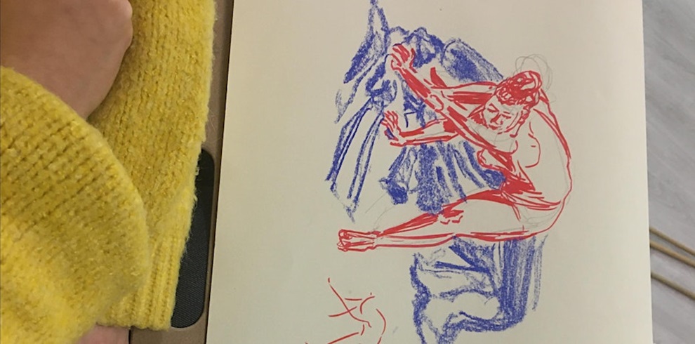 A sketchbook page featuring a life drawing in red and blue. The red line drawing depicts a seated figure with arms outstretched and legs extended, while blue shading suggests draped fabric or background texture. In the foreground, a person wearing a bright yellow knitted sweater rests their arm on the edge of the sketchbook. The scene captures the process of observational figure drawing.