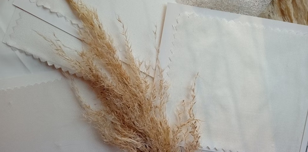 A close-up of handmade paper samples in soft white and cream tones, arranged in overlapping layers with deckled edges. Resting on top is a sprig of dried beige grass, adding a natural, rustic touch to the composition. The scene evokes a minimalist and organic aesthetic, often associated with papermaking or craft workshops.