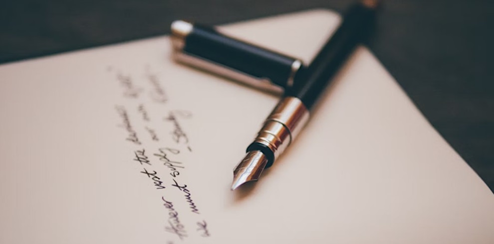 A close-up of a fountain pen resting on a sheet of paper with handwritten cursive text. The pen cap lies beside it, and the focus is on the metallic nib, with the writing slightly blurred in the background.