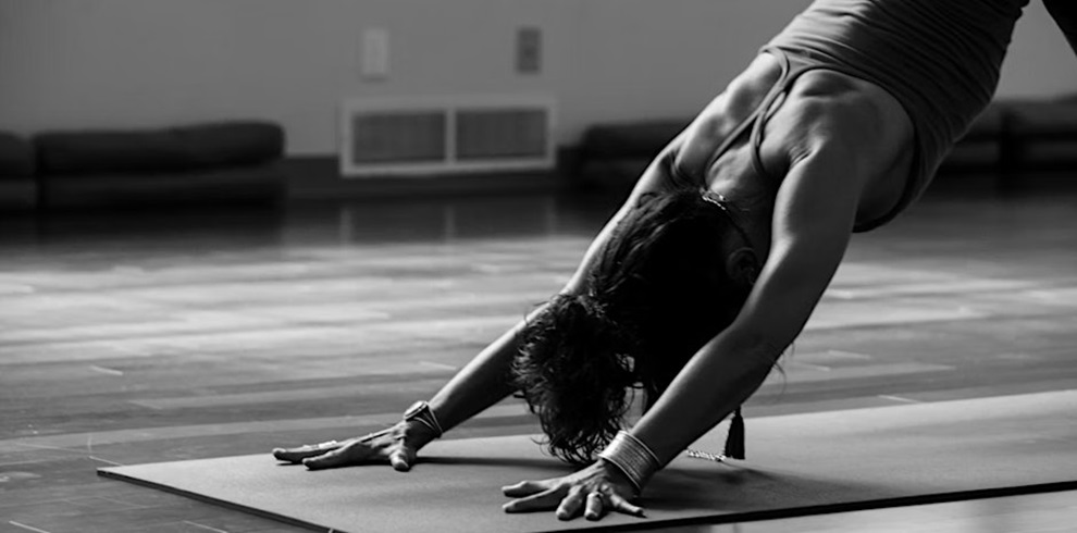 A person practicing yoga indoors, captured in black and white, holding the downward-facing dog pose on a yoga mat with arms stretched forward and back straight.