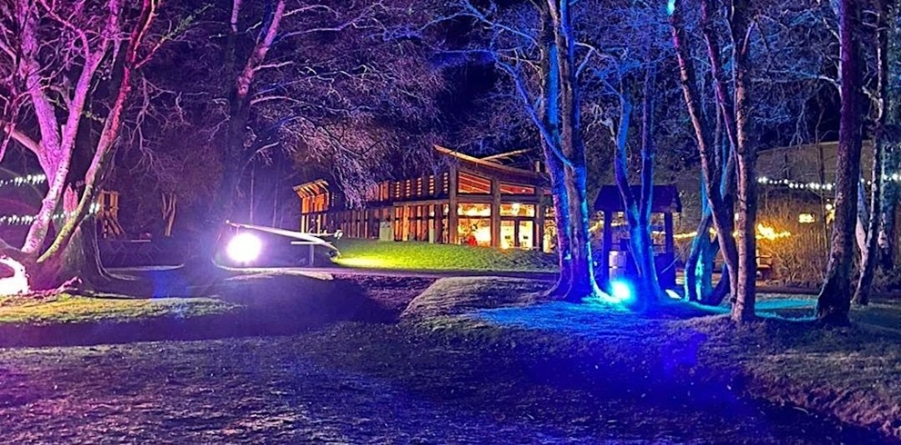 Nighttime scene of a modern wooden building with large glass windows, warmly lit from inside. The structure is surrounded by leafless trees illuminated by vibrant purple and blue lights, creating a colorful glow across the ground. Strings of white lights are visible in the background, adding a festive atmosphere. The foreground shows a grassy and earthy area with patches of light reflecting from the colored illumination.