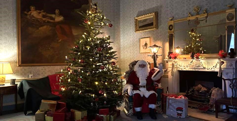A warmly lit living room decorated for Christmas, featuring a large Christmas tree adorned with lights and red ornaments, surrounded by wrapped presents. Santa Claus sits in an armchair beside the tree, waving toward the camera. The room has elegant décor, including a fireplace with garlands and candles, framed paintings on the walls, and a large mirror above the mantel.