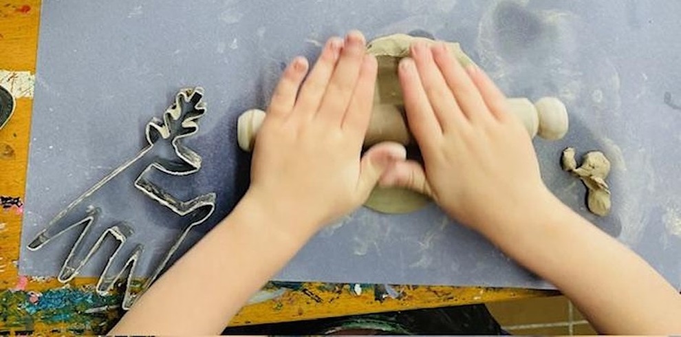 Two small hands pressing down on a piece of clay with a rolling pin on a blue mat. To the left, there is a metal cookie cutter shaped like a reindeer, and to the right, a small piece of clay is rolled into a rough shape. The surface underneath is a wooden table with colorful paint marks.