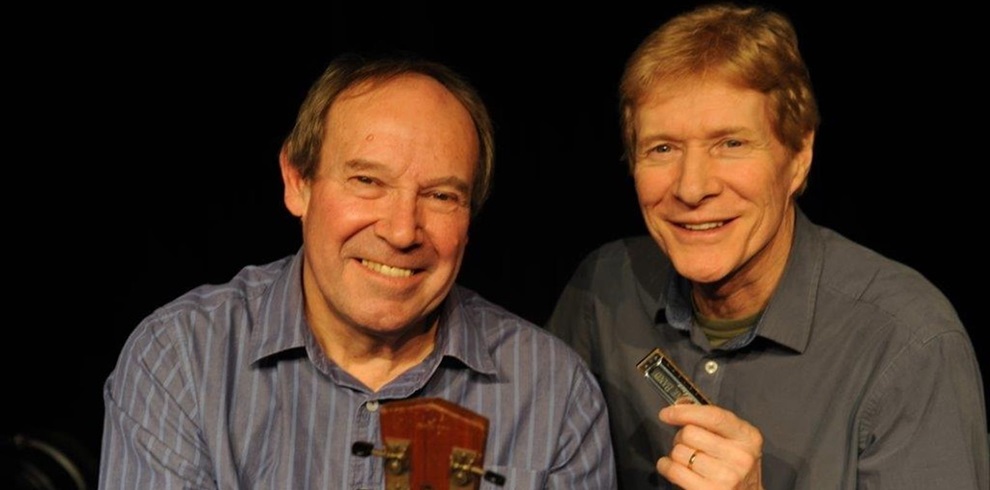 Two musicians are pictured against a dark background. One is holding an acoustic guitar, while the other is holding a harmonica. Both are dressed in casual shirts, and the scene suggests a live performance or promotional photo for a blues or acoustic music event.