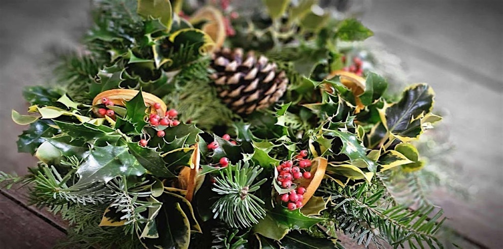 A festive wreath made of natural greenery, including holly leaves, pine branches, and sprigs of red berries. The arrangement also features dried orange slices and a large pinecone in the center, creating a traditional holiday decoration with rich green, red, and brown tones.