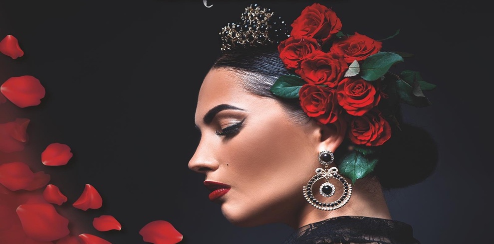 A person wearing an ornate black outfit with intricate lace details is adorned with a decorative headpiece featuring a cluster of vibrant red roses and green leaves. The headpiece also includes a metallic crown-like accessory with small embellishments. Large, circular earrings with black and silver accents hang prominently. On the left side of the image, several scattered red rose petals appear against a dark background, creating a dramatic and elegant composition.