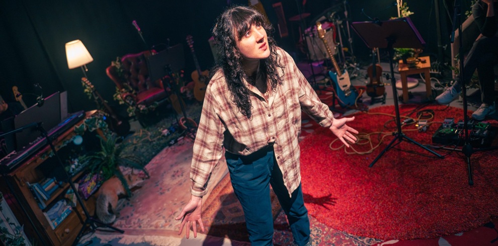 A picture of a person on stage with an orange checked shirt on and jeans.