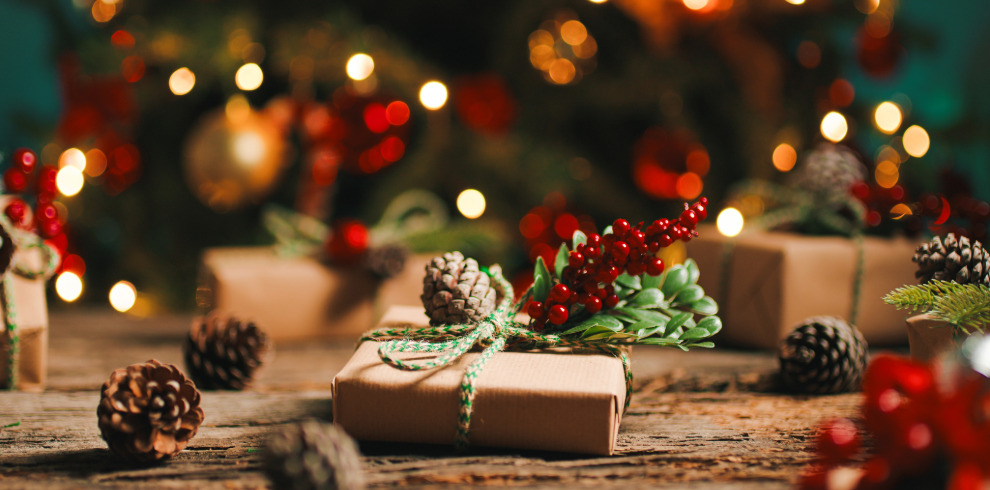 Festive scene with present on table wrapped in brown paper. Tree and lights in background.