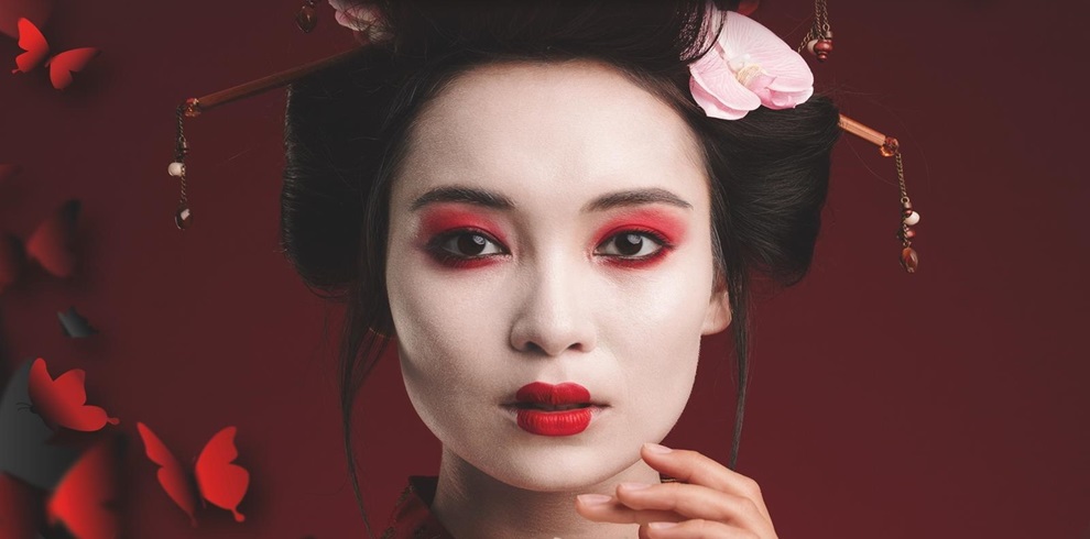 A woman dressed in traditional Japanese-style attire with white face makeup, red eye shadow, and red lipstick poses against a deep red background. Her dark hair is styled up with decorative hairpins and pink flowers. Red butterfly shapes appear along the left side of the image, blending into the background.