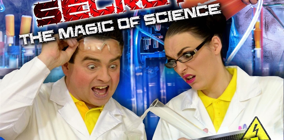 Promotional poster featuring two people wearing white lab coats over bright yellow shirts, standing in front of a colorful science-themed background with test tubes and laboratory equipment. One person is holding a rolled-up paper, and the other is adjusting safety goggles. Bold text at the top reads “SECRET” in red and “THE MAGIC OF SCIENCE” in white, with a yellow caution symbol featuring a lightning bolt at the bottom right.