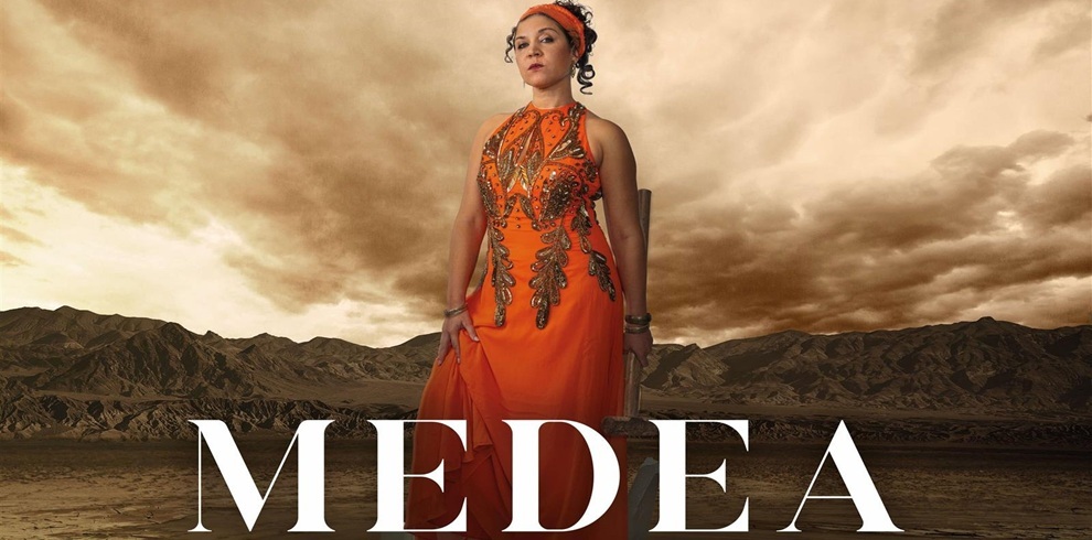 A woman stands confidently in the center of the image against a dramatic, cloudy desert landscape. She is wearing a bright orange, sleeveless dress decorated with intricate gold and bronze beadwork. Her hair is tied back with an orange headband, and she holds a serious, commanding expression. The word “MEDEA” is written in large white capital letters across the bottom of the image.