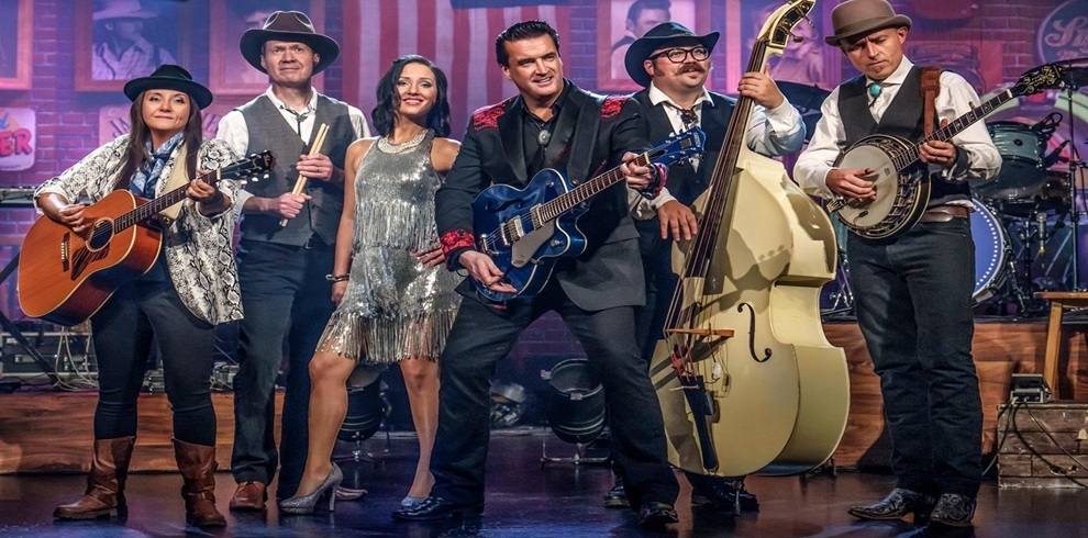 Stage performance featuring a country and rockabilly-style band with six musicians holding instruments including acoustic guitar, electric guitar, upright bass, and banjo. The performers are dressed in vintage-inspired outfits such as cowboy hats, vests, and a silver fringed dress. The background shows a rustic set design with signage and musical equipment.