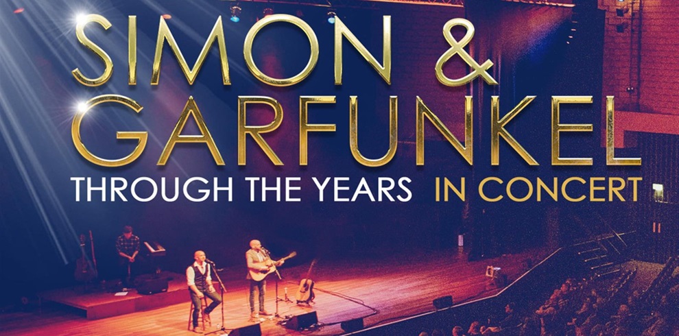 Simon & Garfunkel Through the Years In Concert.” Here’s what the image conveys: Foreground: Two performers on stage with guitars, seated on stools, under warm spotlights. Background: A large concert hall with rows of seats and an audience visible on the right side. Text: Bold gold lettering for “Simon & Garfunkel” and smaller white and gold text for “Through the Years In Concert.” The design clearly communicates that this is a tribute or themed concert celebrating Simon & Garfunkel’s music over the years.