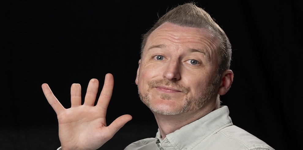 A middle-aged man with short light hair and a trimmed beard is posing against a black background. He is wearing a light gray shirt and raising his left hand with fingers spread, giving a friendly expression with a slight smile.
