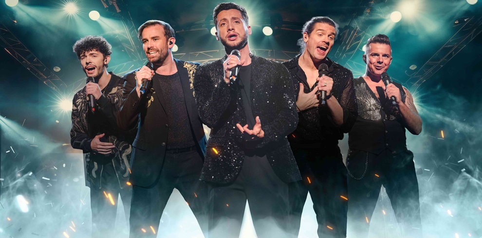 Five male singers performing on stage under dramatic lighting, each holding a microphone and dressed in stylish black outfits with sparkling or patterned details. The background features stage lights, smoke, and an energetic concert atmosphere.