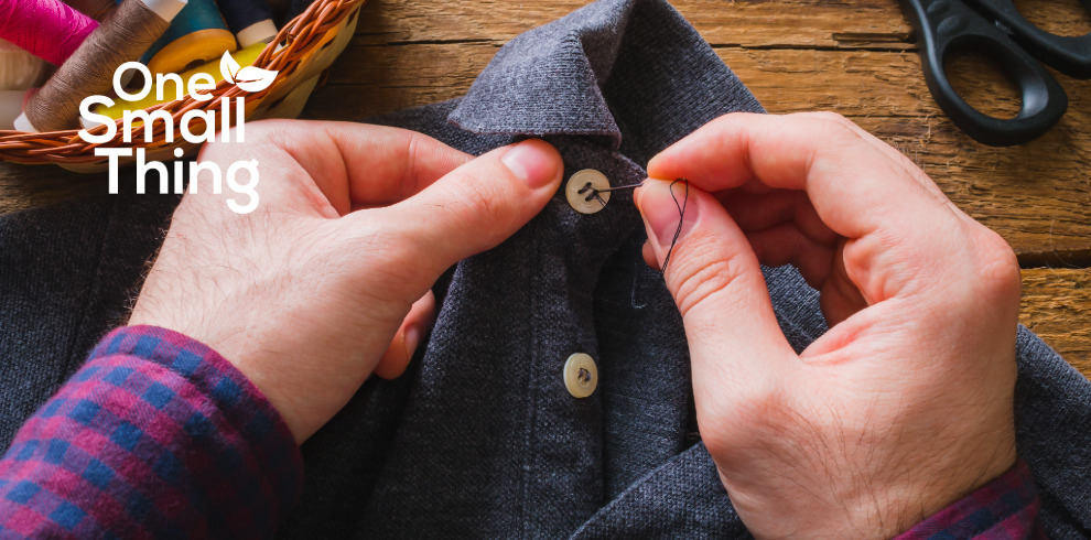 Close-up of hands sewing a button onto a dark grey garment on a wooden table, with thread, scissors, and sewing supplies nearby. The words “One Small Thing” appear in white text in the top left corner.