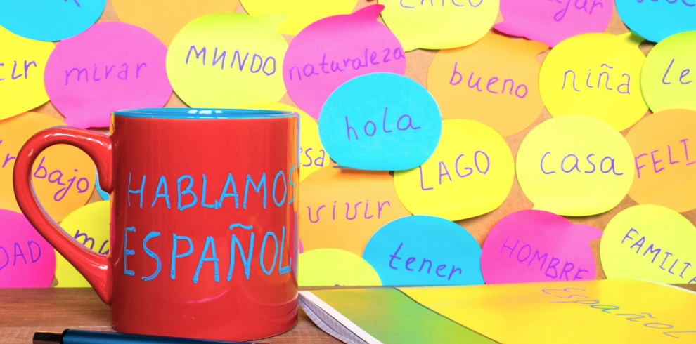 A bright red mug with the phrase “Hablamos Español” written in blue sits on a wooden surface. Behind it, a wall is covered with colourful sticky notes in pink, yellow, blue, and green, each displaying different Spanish words such as “hola,” “mundo,” “casa,” “tener,” and “feliz.” A pen and a Spanish-language notebook are placed in the foreground.