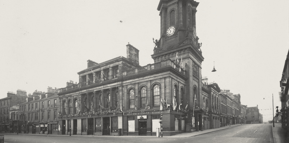 The History of Ayr Town Hall Exhibition 5 Historic urban street scene featuring a classical-style building with a clock tower reading 11:50. The structure displays arched windows, ornate architectural details, and signage reading "HUNTER & GRANT" and "Establishment." Flags and banners suggest a celebratory event. Surrounding buildings reflect early 20th-century design, and the street is empty of people and vehicles.