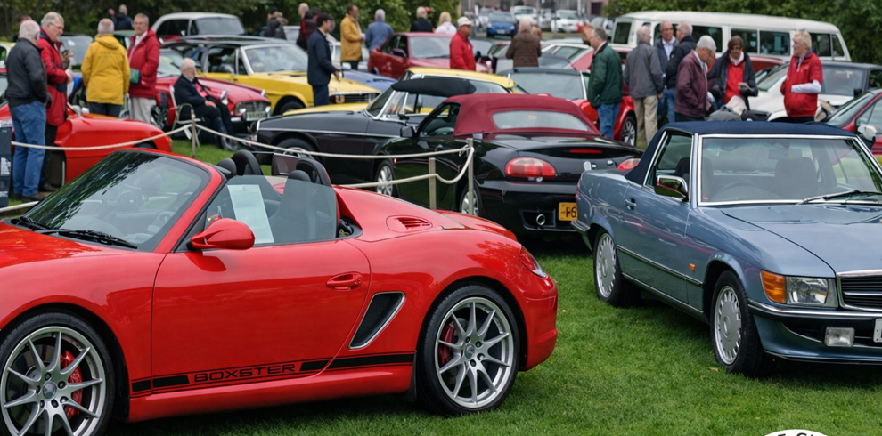 A crowded classic car show on a grassy field, featuring rows of vintage and modern convertibles. In the foreground, a bright red Porsche Boxster with the top down is parked beside a blue Mercedes-Benz convertible. Behind them, various classic cars in black, yellow, and red are roped off while a group of mostly older adults walk around, chatting and admiring the vehicles.