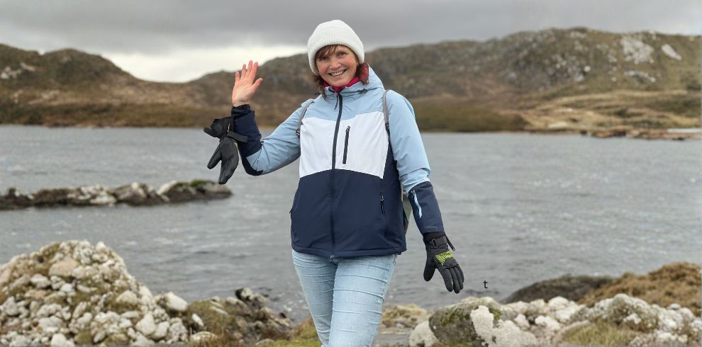 A woman smiling and waving at the camera on a beach.