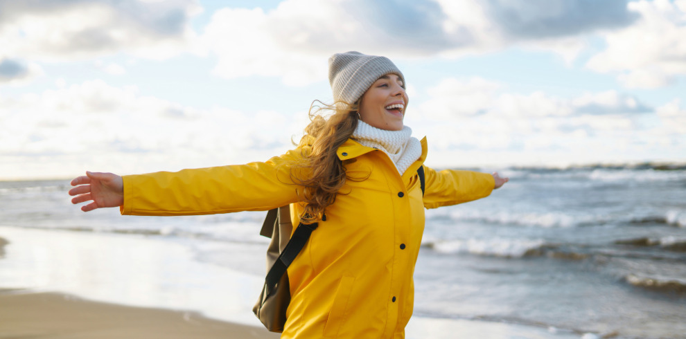 Smiling woman in a yellow jacket with arms outstretched, standing on a windy beach in winter, wearing a hat and scarf with the sea behind her