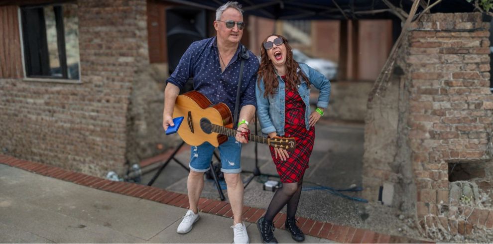 Two people stand side by side outdoors in front of a brick wall. One holds an acoustic guitar while the other rests a hand on the guitar’s neck. They appear to be posing together under a canopy, with various cables and equipment visible on the ground behind them.