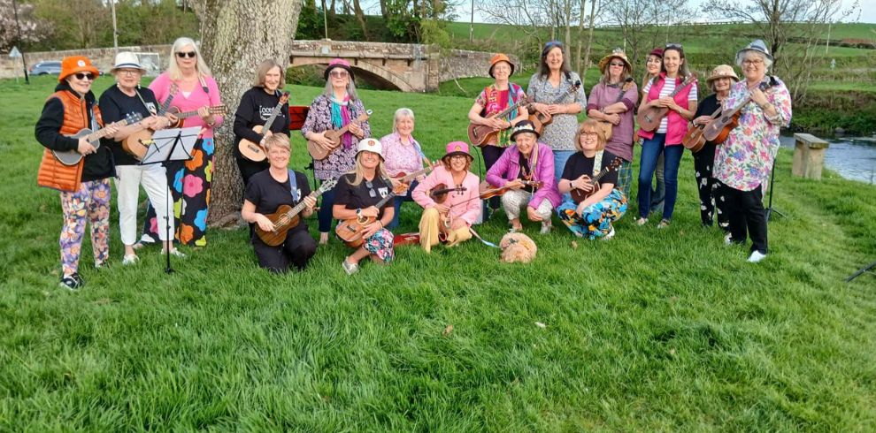 A group of people are gathered outdoors on a grassy area beside a tree and a small river. They are standing and sitting in a semi‑circle, each holding a ukulele. The group is dressed in colourful, patterned clothing, and music stands are set up nearby. In the background is a stone bridge, trees, and open green space.