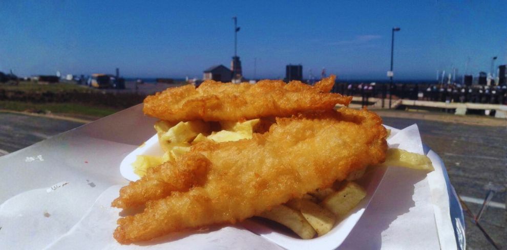 Freshly prepared fish and chips served in a paper tray with a clear blue sky and Girvan Harbour in the background.