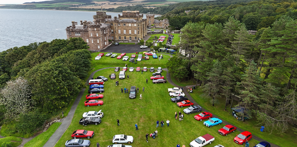 An aerial shot of a large grassy area on the grounds of an historic castle beside the coastline. Dozens of classic cars in various colors are arranged in rows on the lawn, with small groups of people walking among them. Dense trees and views of the sea surround the venue.