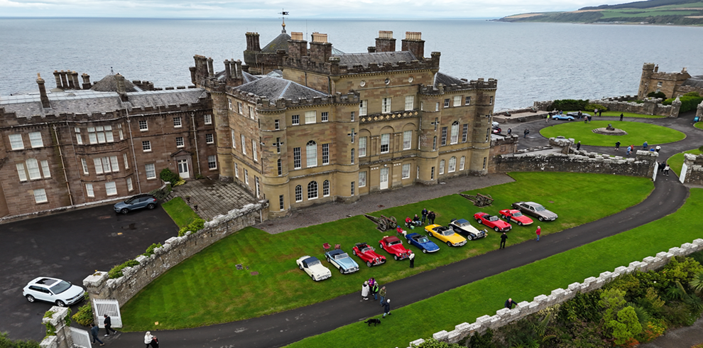 An aerial shot of a large grassy area on the grounds of an historic castle beside the coastline. Dozens of classic cars in various colors are arranged in rows on the lawn, with small groups of people walking among them. Dense trees and views of the sea surround the venue.