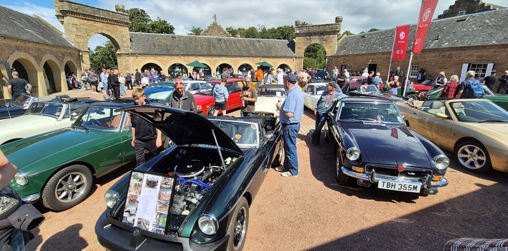 An outdoor classic car show set in a stone courtyard with arched buildings in the background. Many people are gathered around various classic cars, including convertibles and coupes. One dark green car in the foreground has its hood open for display, showing the engine. The weather is sunny with scattered clouds.