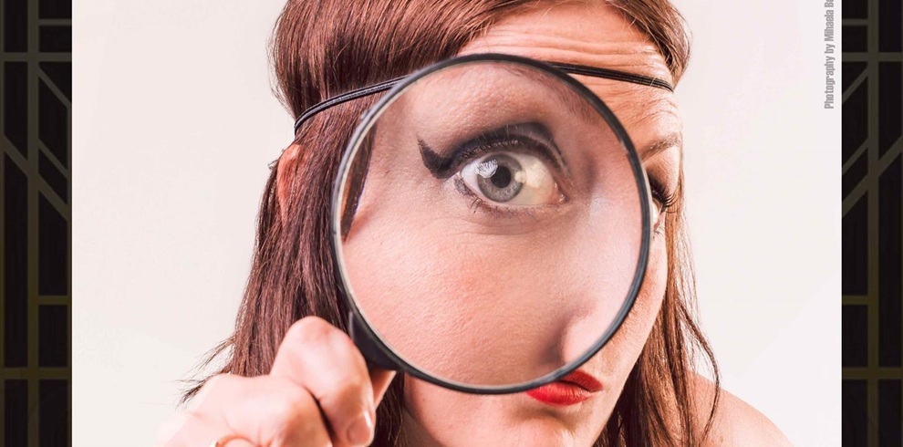 A person with shoulder‑length brown hair holds a magnifying glass toward the camera, creating a close‑up, detective‑themed composition. The background is light, and the scene has a mystery or investigation aesthetic.