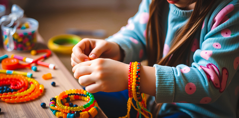 A person with long hair is sitting at a table and threading colorful beads. Several completed beaded bracelets and loose beads are spread across the wooden surface. The person is wearing a light blue top with a pink circular pattern and has multiple bright bead bracelets on their wrist.