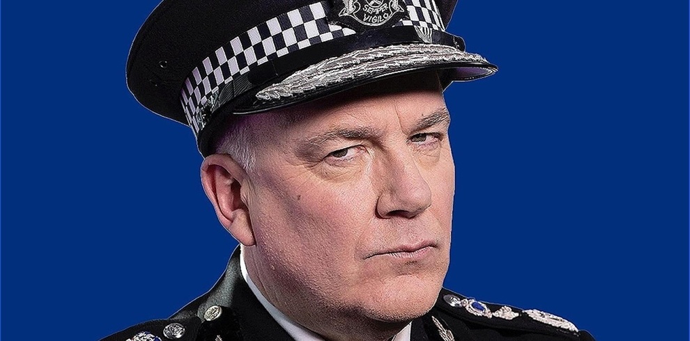 A person wearing a formal police uniform and peaked cap with a black‑and‑white checked band stands against a solid blue background. The uniform includes decorative silver detailing and insignia.