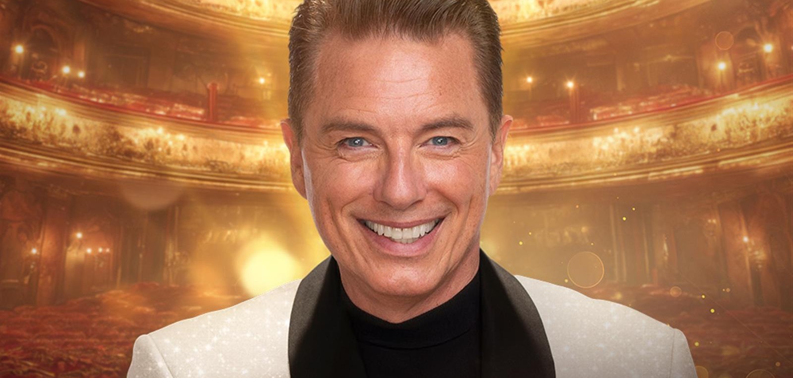 A person wearing a light-coloured suit jacket with a black lapel stands in front of a grand, warmly lit theatre interior. The background features ornate balconies, golden lighting and an elegant auditorium, creating a glamorous, theatrical atmosphere