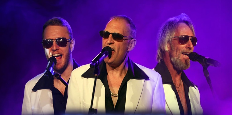 Three performers dressed in white jackets with black collars stand in front of microphones on a stage filled with purple and blue concert lighting. They appear to be singing as part of a music tribute show.