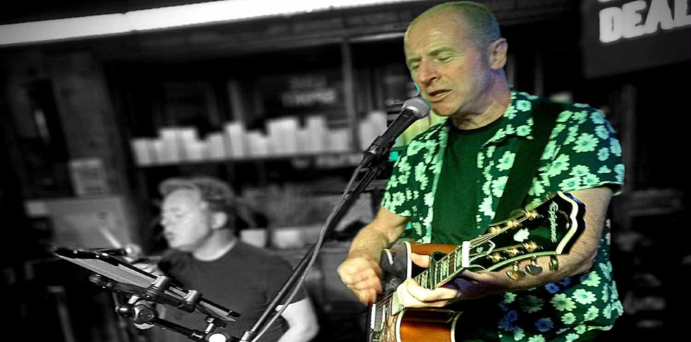 A person in a floral-patterned shirt stands at a microphone while playing an electric guitar on stage. Another person sits nearby behind a music stand with a tablet or sheet holder. The background shows shelves and signs in a dimly lit indoor venue, with selective color highlighting the performer in the foreground.