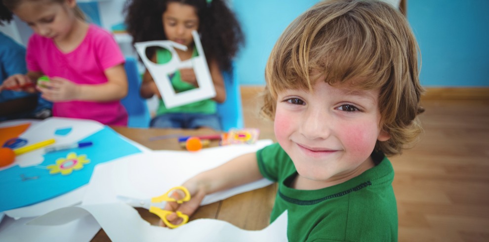 Children sitting at a table engaged in an arts and crafts activity, using paper, scissors, and colorful materials in a bright classroom setting