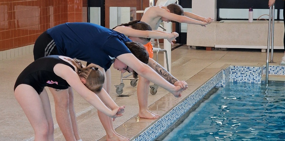 Row of children getting in a diving pose next to a swimming pool.