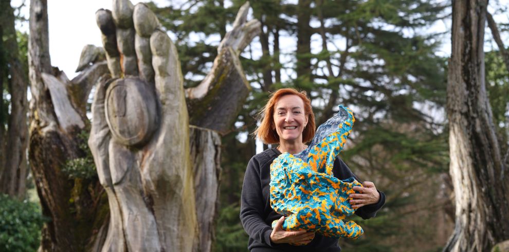A person with short, light-colored hair stands outdoors in front of a large wooden sculpture shaped like an upright hand with carved fingers. The person is holding a brightly colored abstract object featuring blue, yellow, and orange patterns. Tall trees fill the background, and the scene appears to be in a natural or park-like setting.