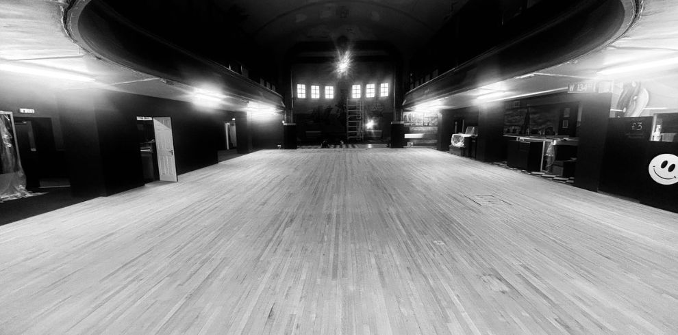 A black‑and‑white photo of a large empty dance hall with wooden flooring, curved ceiling.